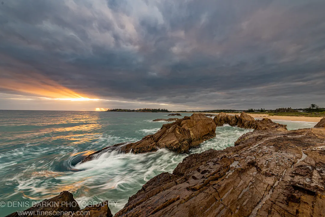Long-exposure wave-wash over layered rock platforms at Bermagui, with golden light breaking through storm clouds at dawn.