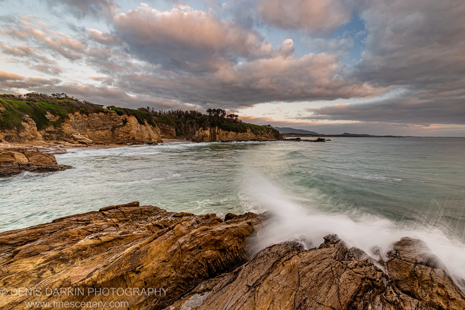 Sandstone rocks and breaking waves in the foreground, with a scrub-covered headland curving into a calm turquoise bay at dawn.