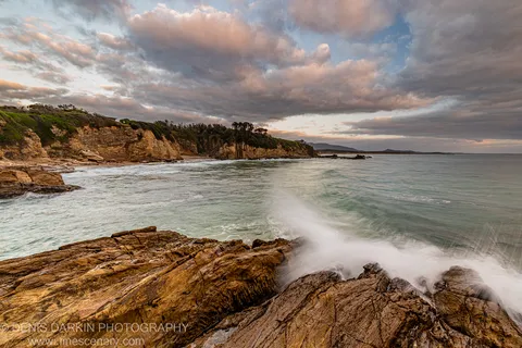 Sandstone rocks and breaking waves in the foreground, with a scrub-covered headland curving into a calm turquoise bay at dawn.