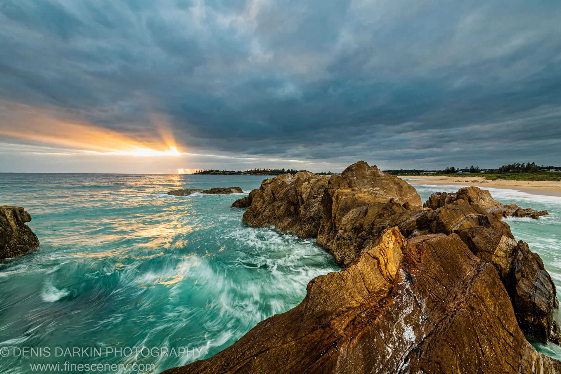 Golden-brown sandstone on the NSW South Coast lit by a shaft of sun breaking through heavy cloud, with shimmering reflections on the waves below.