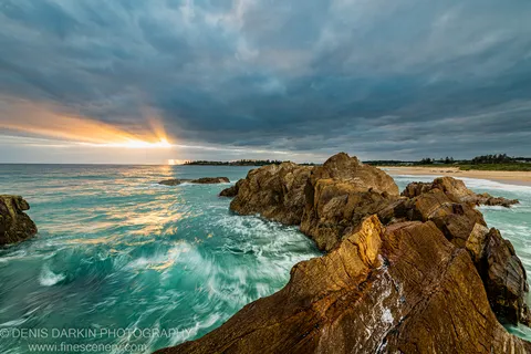 Golden-brown sandstone on the NSW South Coast lit by a shaft of sun breaking through heavy cloud, with shimmering reflections on the waves below.