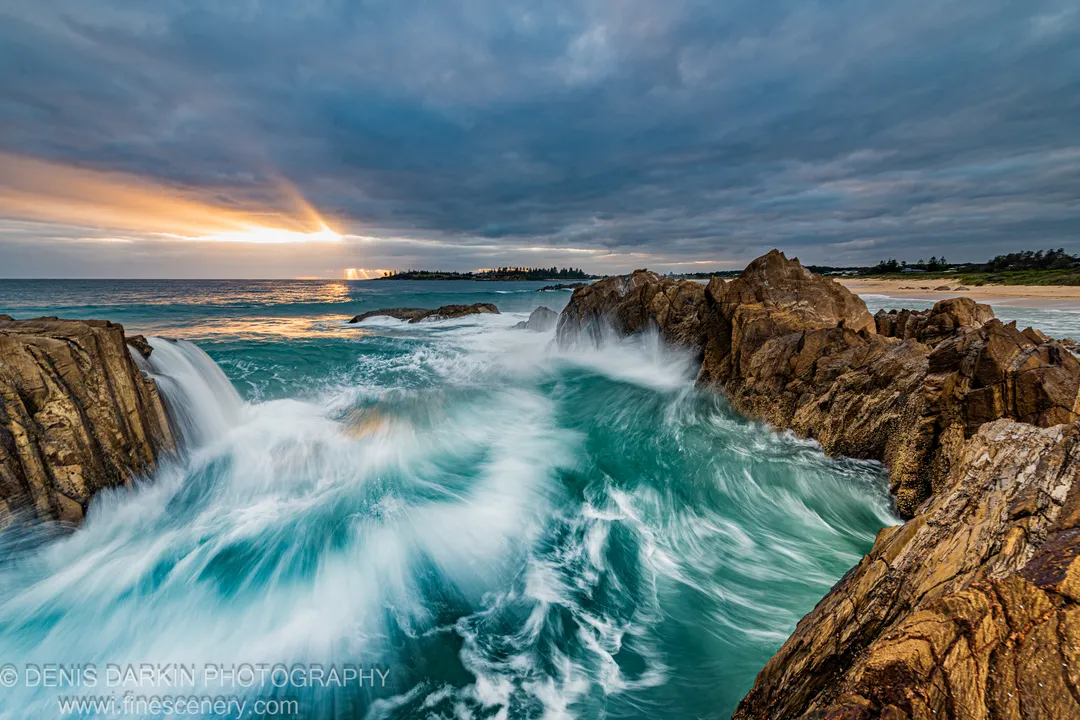 A bright sunburst breaks through heavy storm clouds over the South Coast of NSW, casting warm light across the ocean below at dawn.
