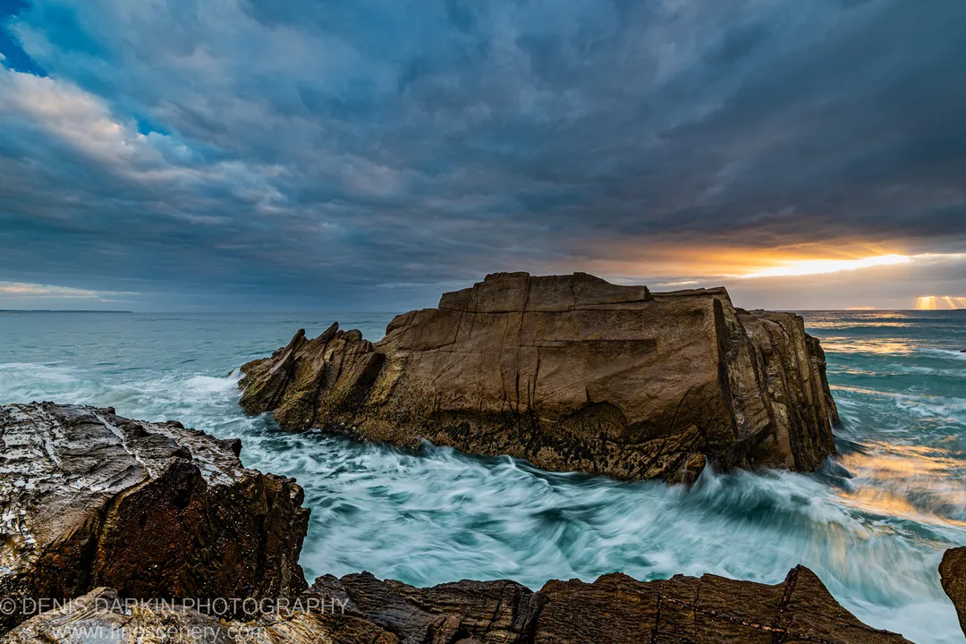 A sandstone headland juts into the ocean on the NSW South Coast, golden sunrise rays breaking through storm cloud and turquoise waves at the base.