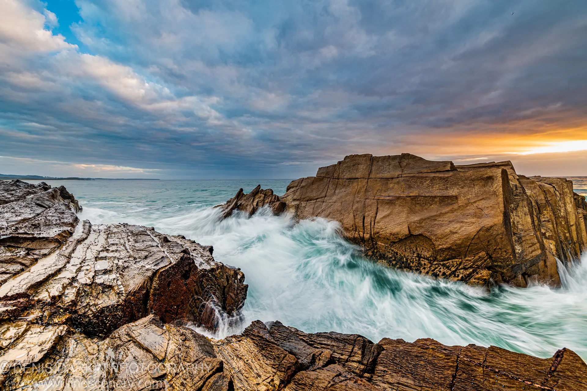 Rugged rocky shore with white-capped waves below a large sandstone cliff on the South Coast of New South Wales at dawn.