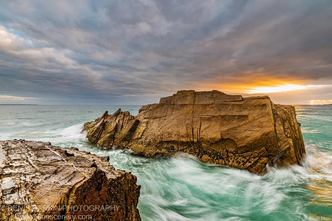 Massive stratified sea stack rising from the ocean on the South Coast of NSW at dawn with storm clouds and a warm sunrise glow.