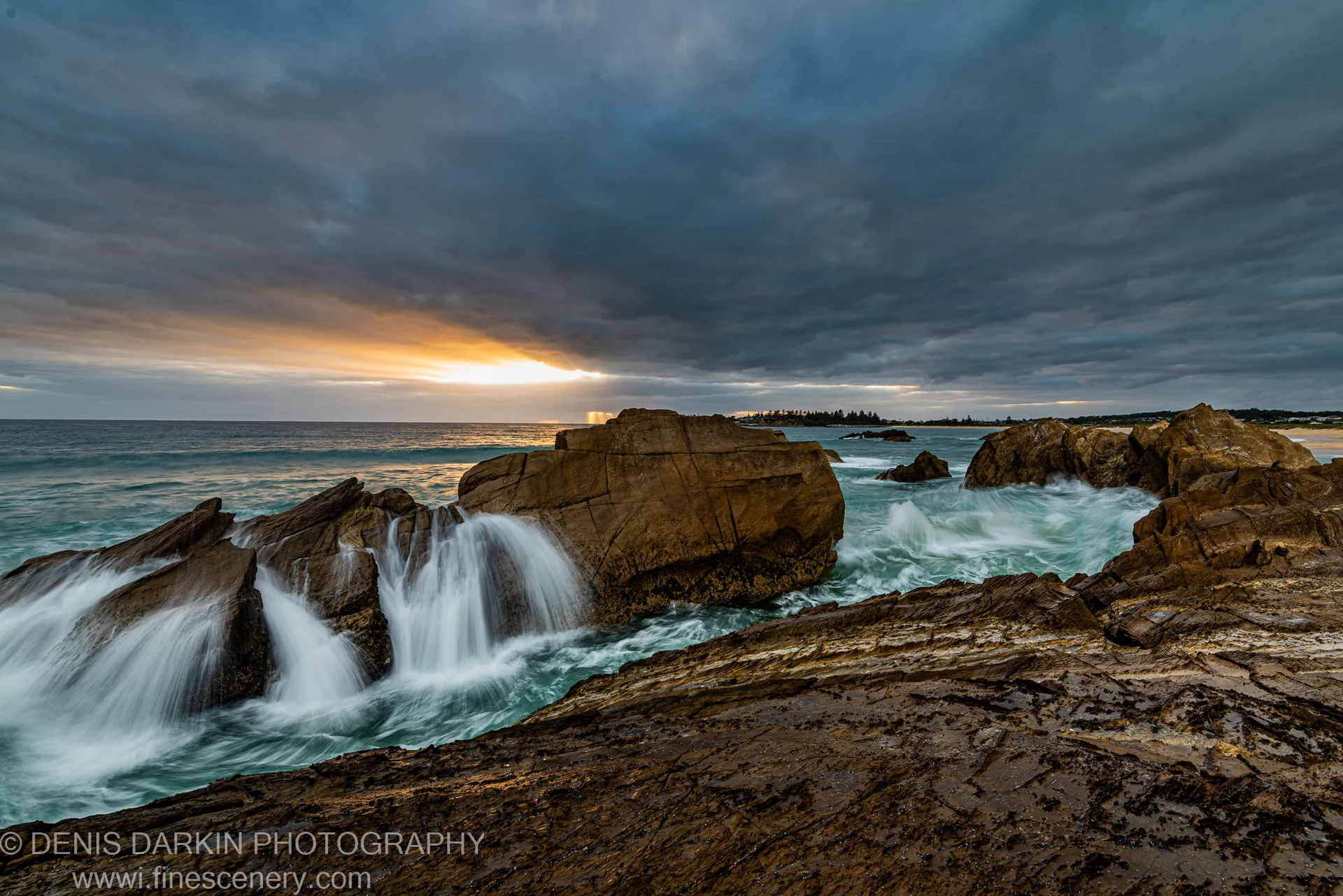 Amber light on the horizon breaking through storm clouds over a South Coast rock platform with long-exposure water at dawn.