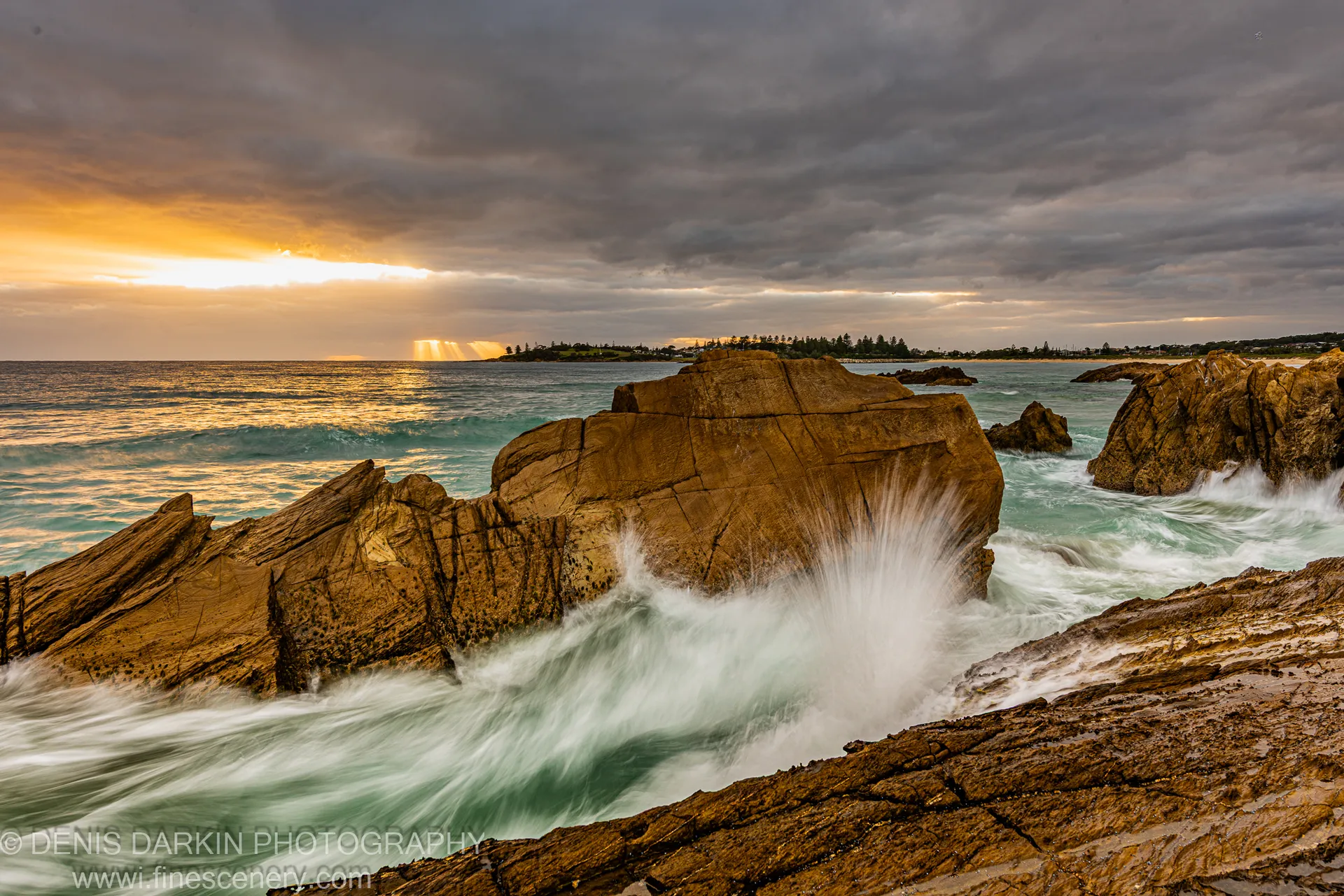Crepuscular rays through cloud breaking over sandstone boulders and turquoise surf on the South Coast of NSW at dawn.