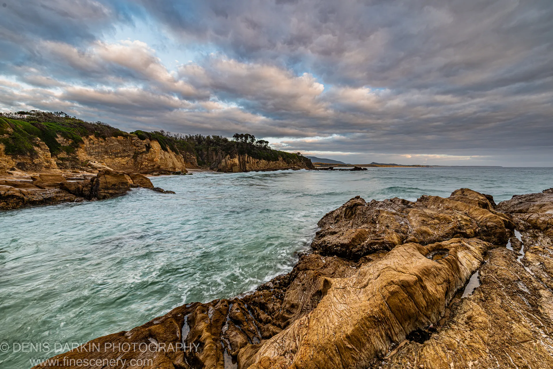 Wet striated sandstone rocks in the foreground with turquoise water and a distant headland on the South Coast of NSW at dawn.