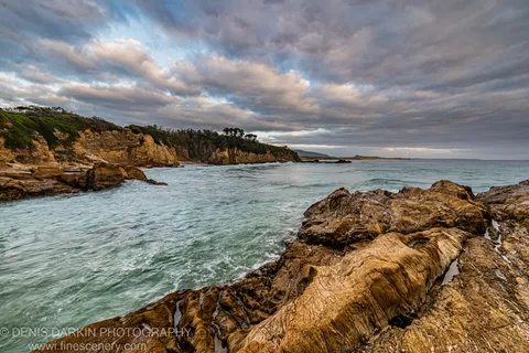 Wet striated sandstone rocks in the foreground with turquoise water and a distant headland on the South Coast of NSW at dawn.
