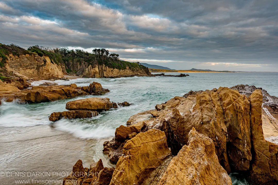 Warm-toned sandstone formations with motion-blurred tidal wash on the South Coast of NSW at dawn, cliffs and headland behind.