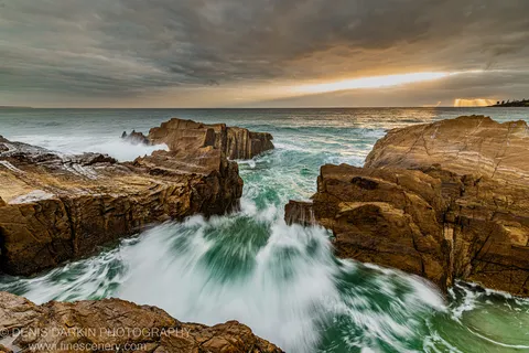 Waves rush through a narrow sandstone gap at dawn on the South Coast, with crepuscular rays in the cloud-filled sky above.