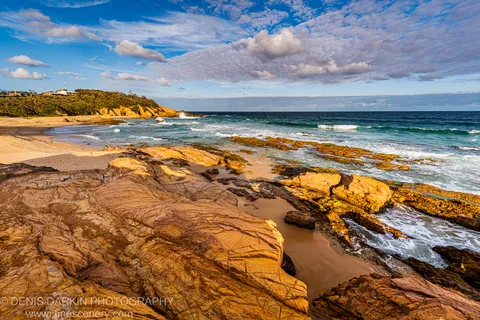 Veined sandstone platform stretches toward a sandy cove at golden hour, waves breaking against a coastal reef under a cloud-scattered sky, New South Wales.