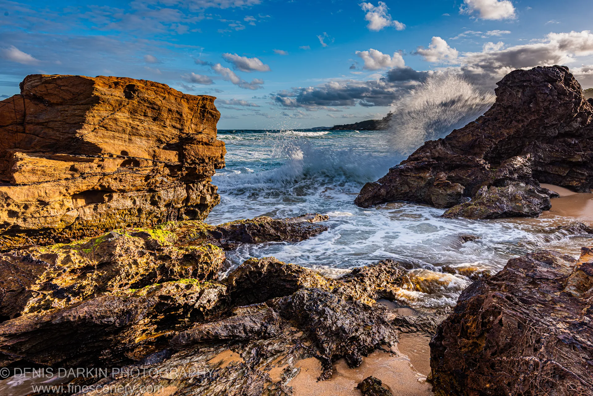 A wave explodes in a white arc against a sandstone cliff at dusk on the South Coast, warm light on the rock face.