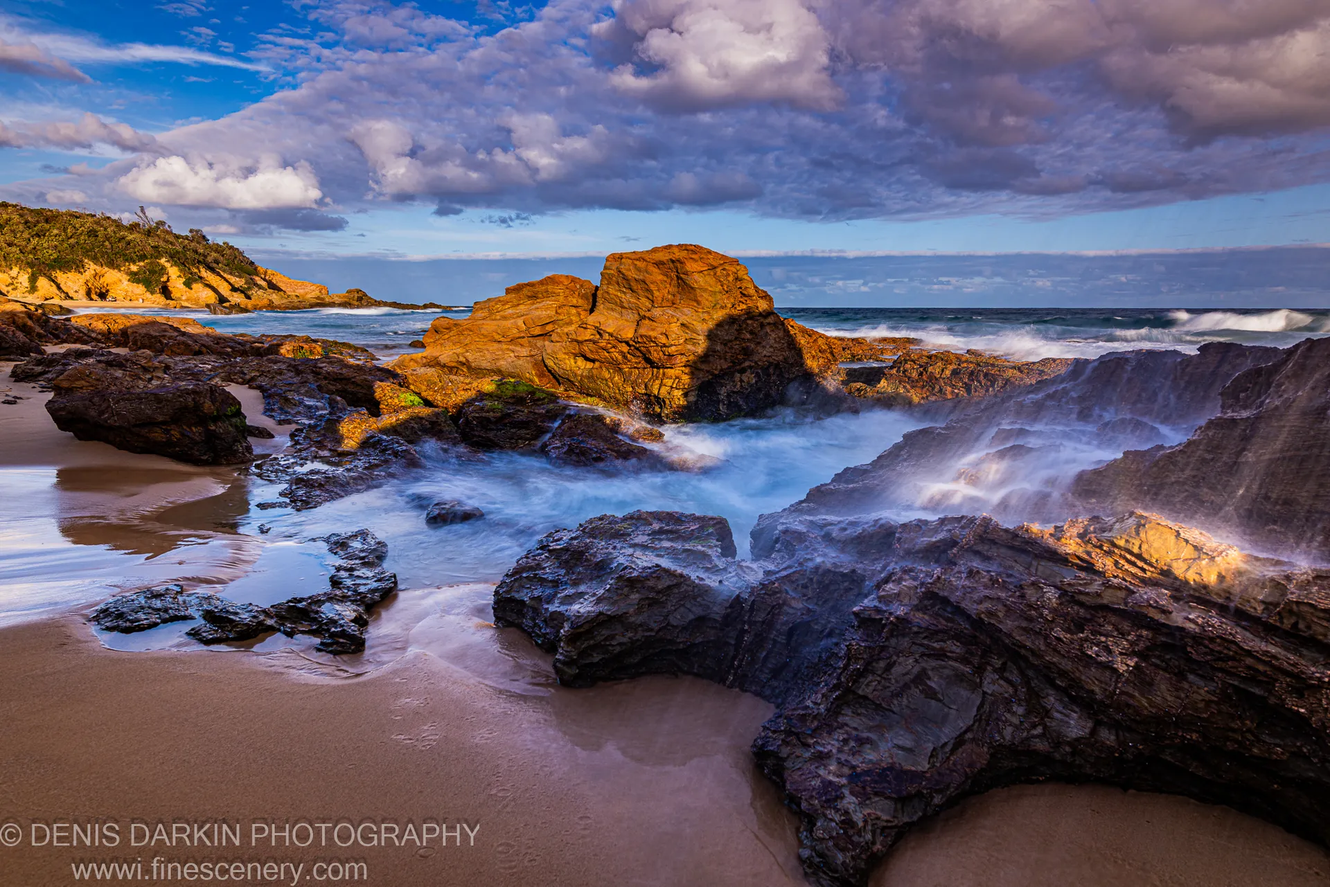 Dark sandstone boulders sit on wet sand at dusk, with long-exposure waves as white mist and a deep blue sky, South Coast NSW.