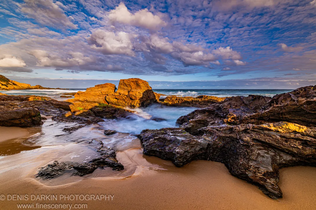Warm afternoon light strikes coastal boulders as smooth, milky water flows over dark stone onto wet sand, with cumulus clouds above the South Coast shoreline.
