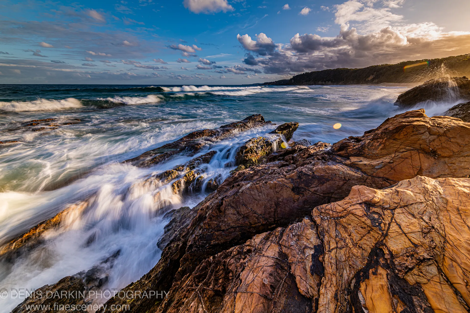 Sun flare marks low dusk light over jagged sandstone rocks, with long-exposure water streaming to the base of a dark headland, South Coast NSW.