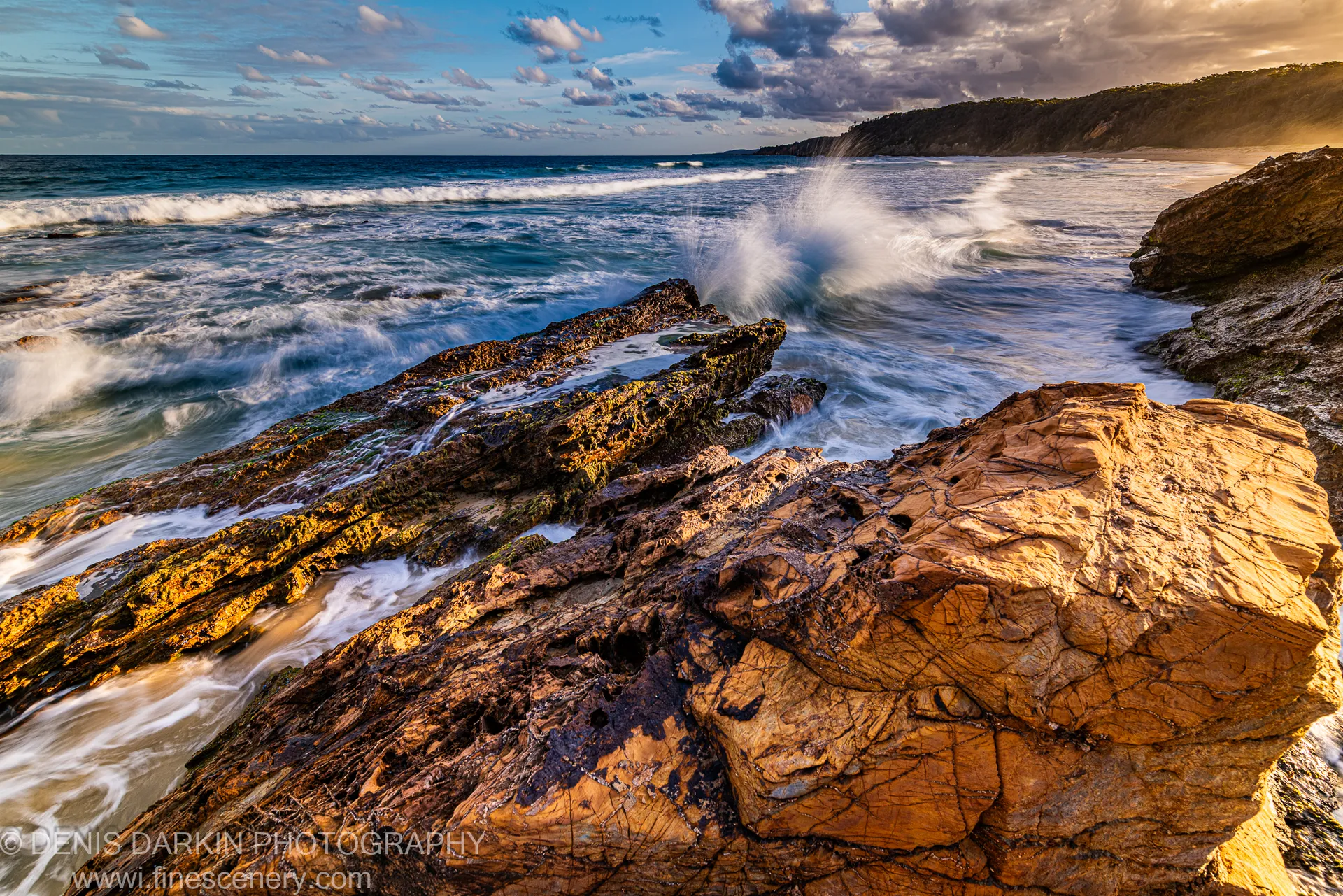A wave crashes against a sandstone platform at dusk, sending a white spray arc against the deep blue sky and distant headland, South Coast NSW.