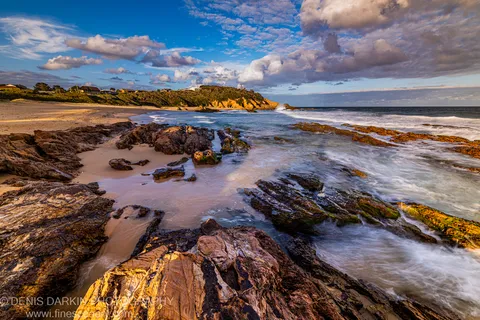 Smooth long-exposure ocean water flows over algae-covered sandstone ledges at dusk, with a sandy cove in the distance, South Coast NSW.