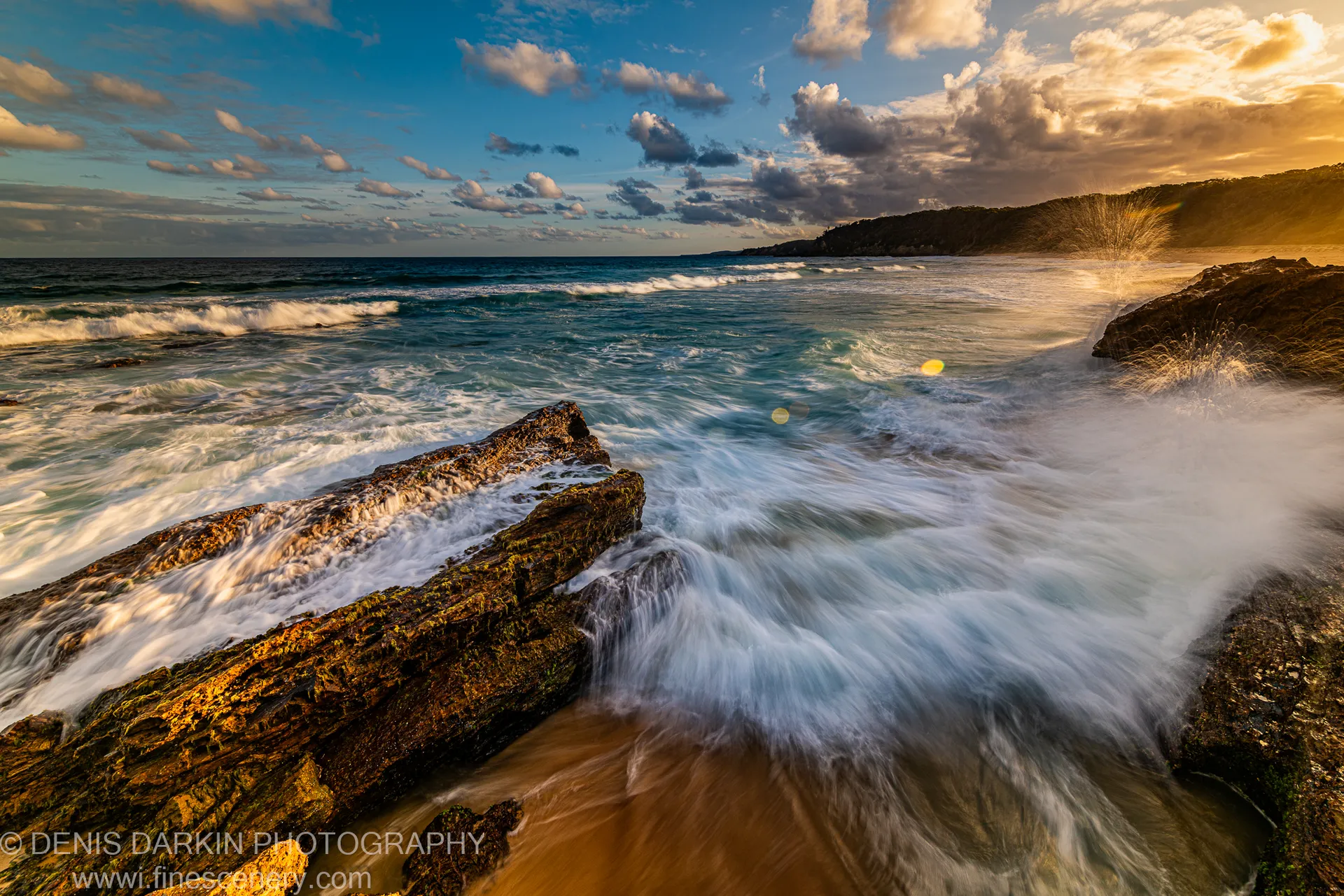 Diagonal volcanic rock platform on the New South Wales South Coast at dusk, with circular lens flare and distant headland beyond.