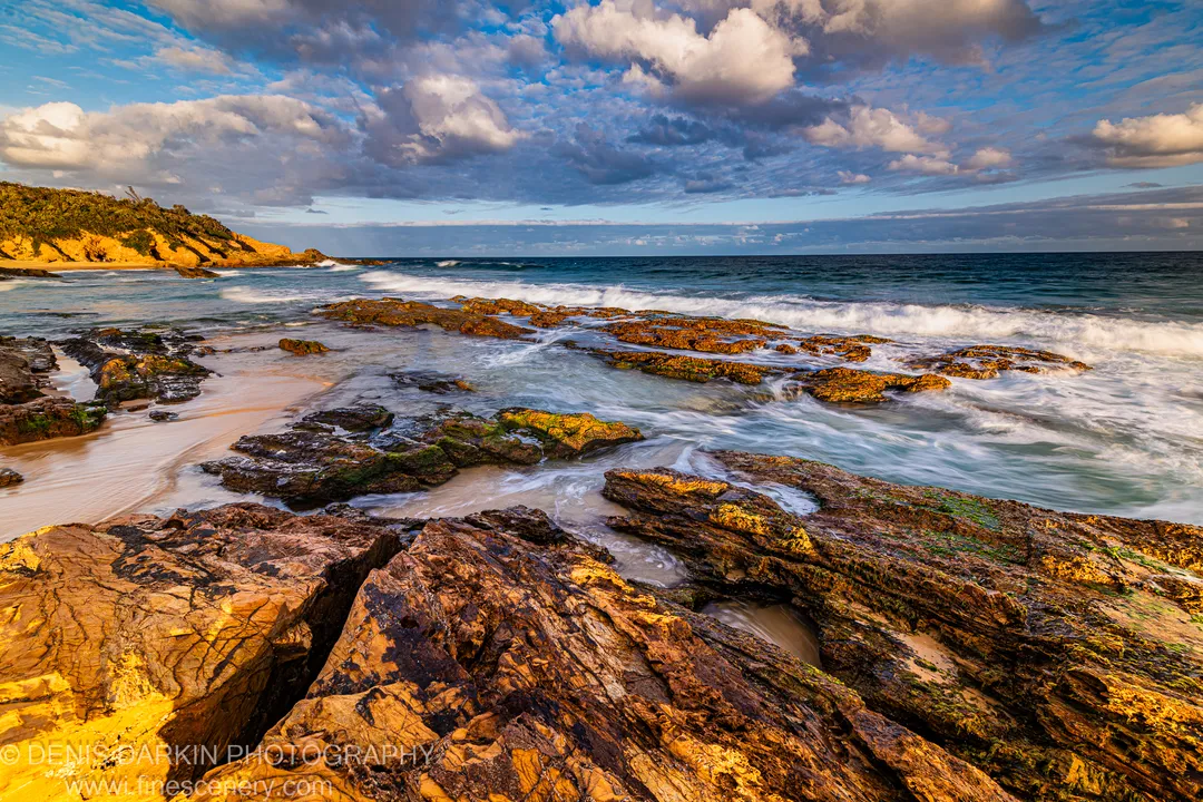 Limestone ledge above a wave-washed sandy cove on the New South Wales South Coast, with motion-blurred seafoam and a distant headland at dusk.