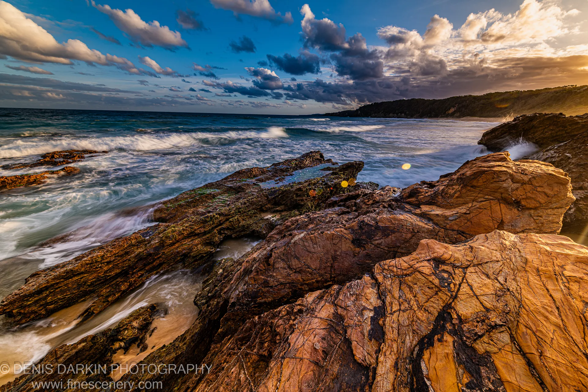 Sandstone coastal shelf in warm dusk light on the New South Wales coast, with breaking waves below and cumulus and storm clouds overhead.