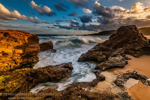 Sea foam between dark boulders below a sandstone cliff face on the New South Wales coast, clouds overhead at dusk.