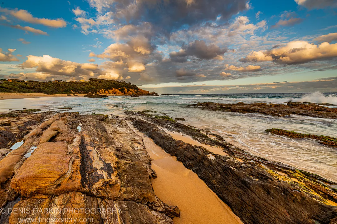 Layered sandstone formations in the foreground lead toward rolling surf and a cliffside residential headland on the New South Wales South Coast at dusk.