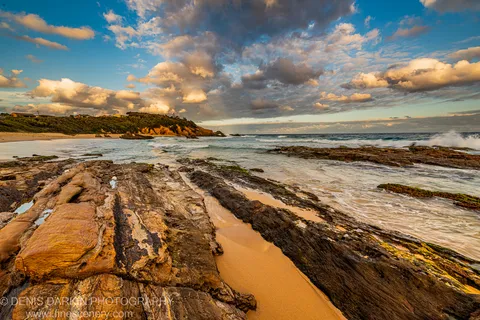 Layered sandstone formations in the foreground lead toward rolling surf and a cliffside residential headland on the New South Wales South Coast at dusk.