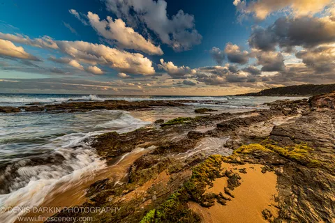 Warm dusk light catches a sandstone platform and algae-covered rocks as cumulus clouds build above a darkened coastal headland, South Coast, New South Wales.