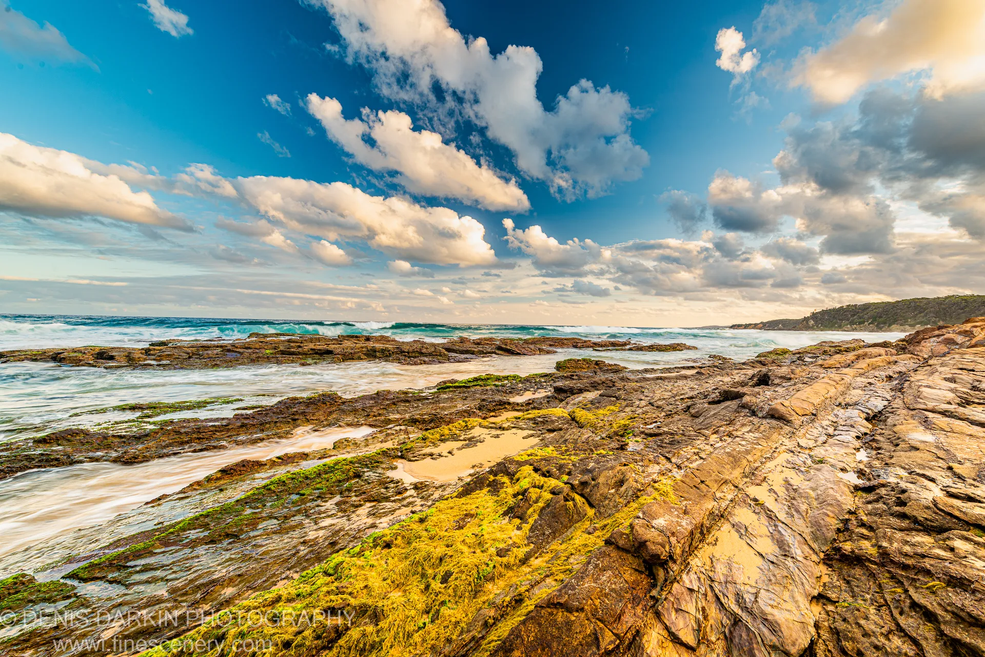Wet, green algae-covered slate rock on the wave-cut platform leads toward breaking surf below backlit cumulus clouds, South Coast, New South Wales.