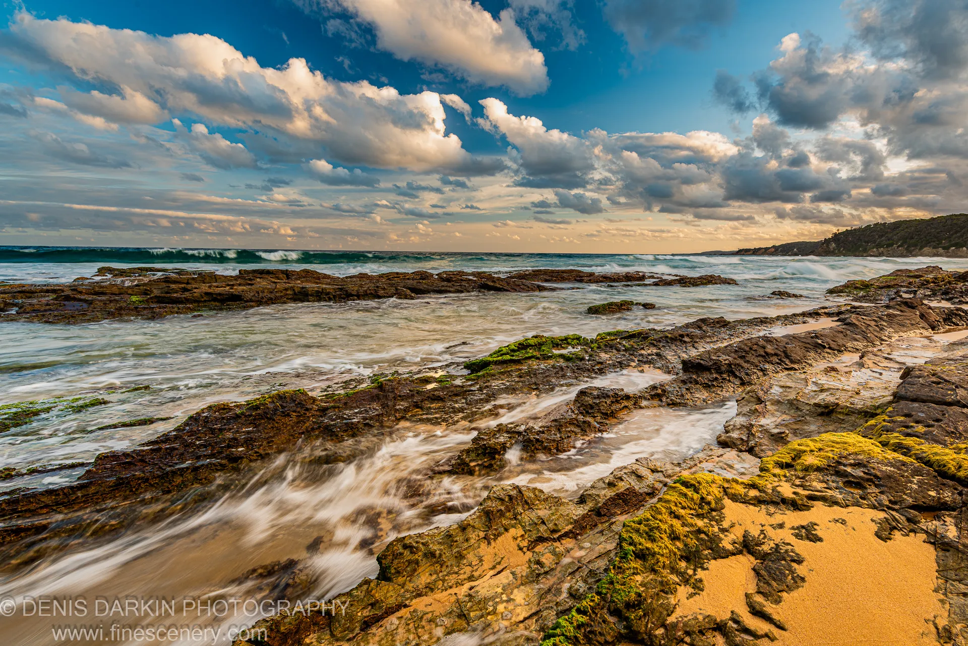 Long-exposure water softens the tidal wash across dark, algae-spotted rock as warm dusk light falls along the horizon, South Coast, New South Wales.