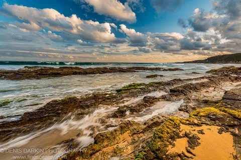 Long-exposure water softens the tidal wash across dark, algae-spotted rock as warm dusk light falls along the horizon, South Coast, New South Wales.