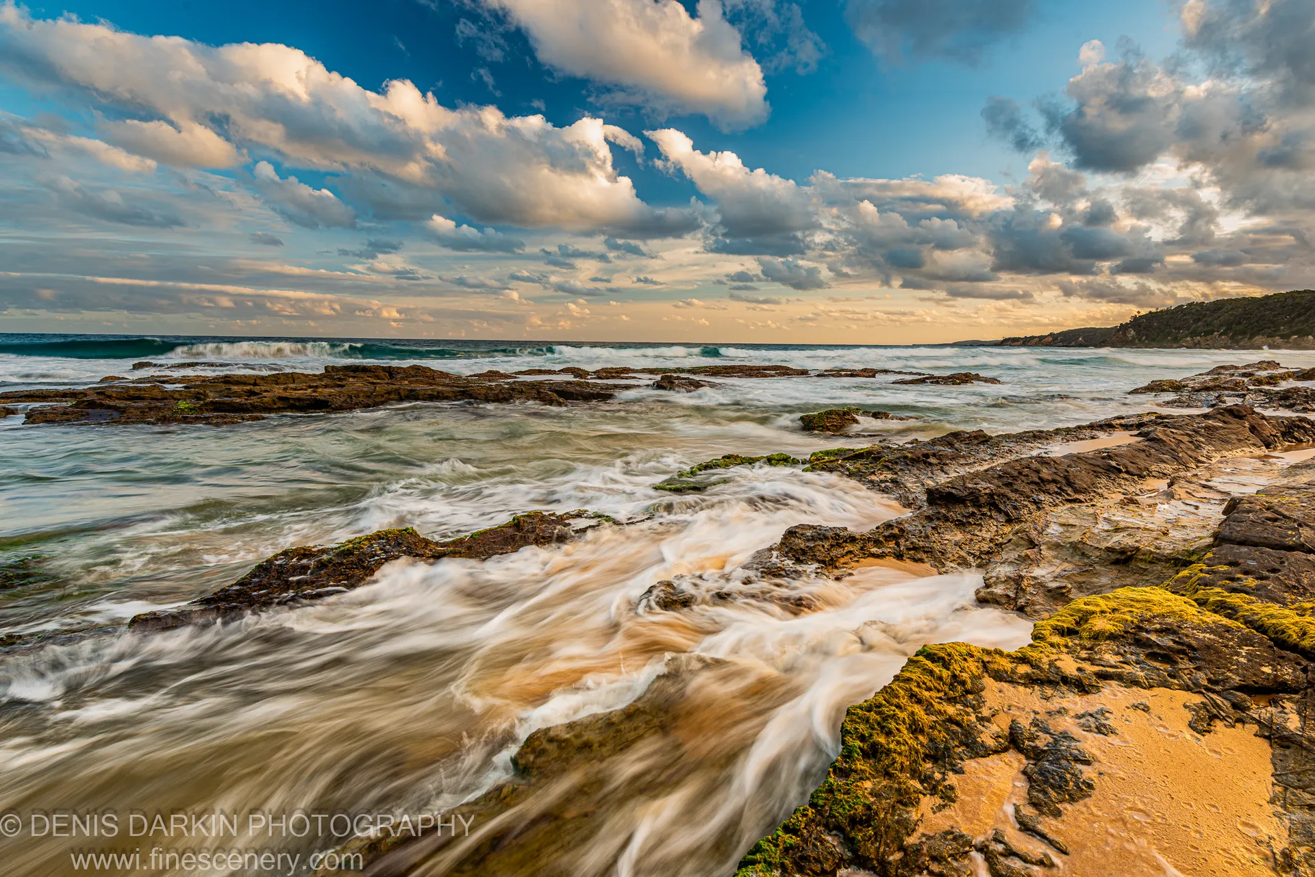 Smooth wave-wash flows over moss-covered rocks on a tidal platform as cumulus clouds fill a deep blue sky above the South Coast, New South Wales.