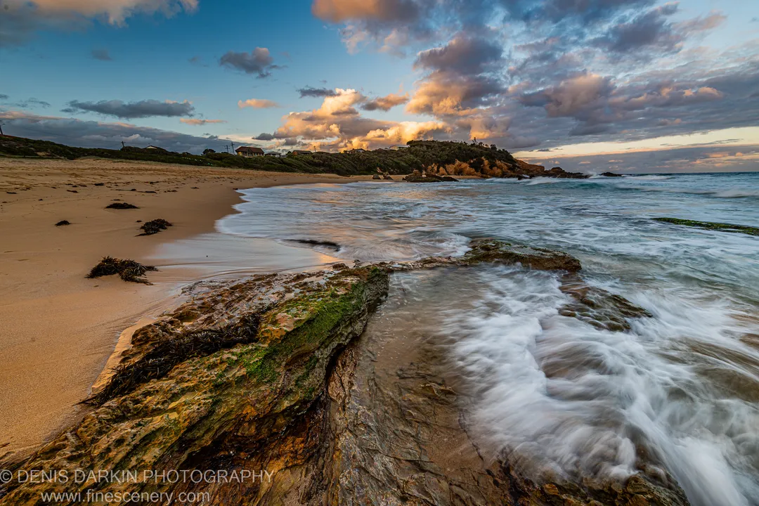 Moss-covered rock in the foreground catches warm dusk light as blurred surf runs past toward a headland with houses, New South Wales.