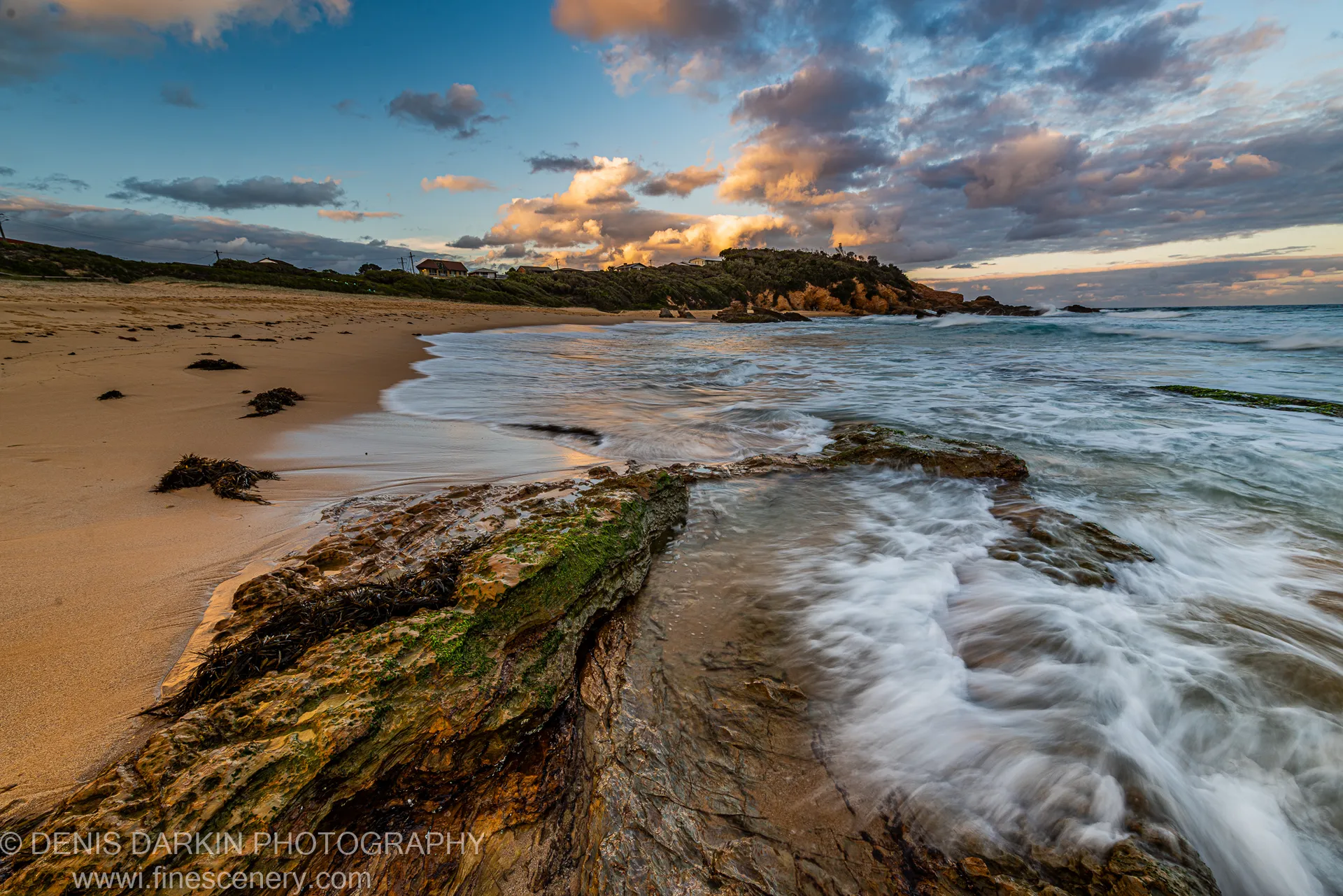 Moss-covered rock in the foreground catches warm dusk light as blurred surf runs past toward a headland with houses, New South Wales.