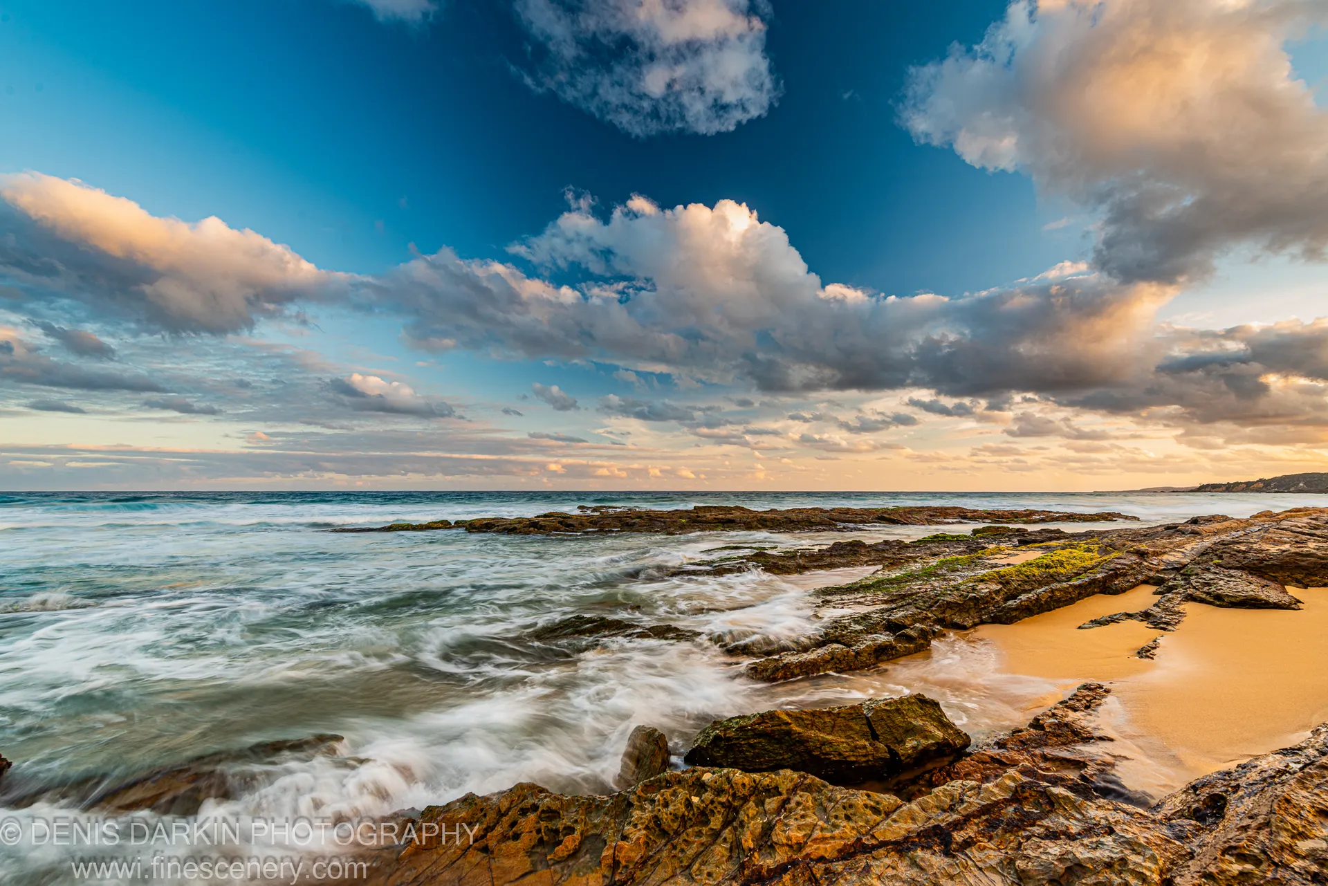 Moss-covered rocks lead toward a sandy inlet where white water moves at dusk, below warm-edged cumulus clouds, New South Wales coast.