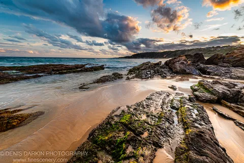 Heavy blue-grey storm clouds contrast with warm golden light along the horizon and a distant rocky headland at evening, South Coast, New South Wales.