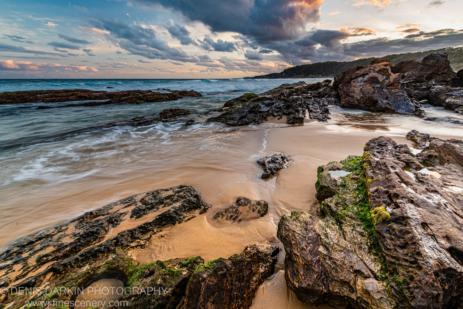 Algae-covered volcanic boulders in the foreground with long-exposure surf and wet sand on the South Coast at evening, New South Wales.