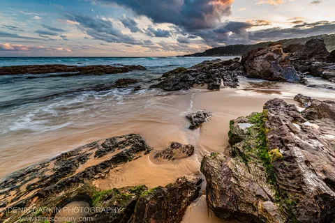 Algae-covered volcanic boulders in the foreground with long-exposure surf and wet sand on the South Coast at evening, New South Wales.