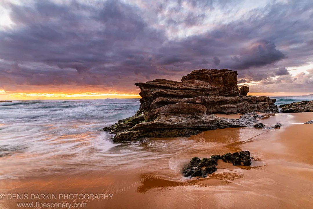 A stratified sandstone rock stack on the South Coast at dawn, with storm clouds above and long-exposure surf on wet sand.