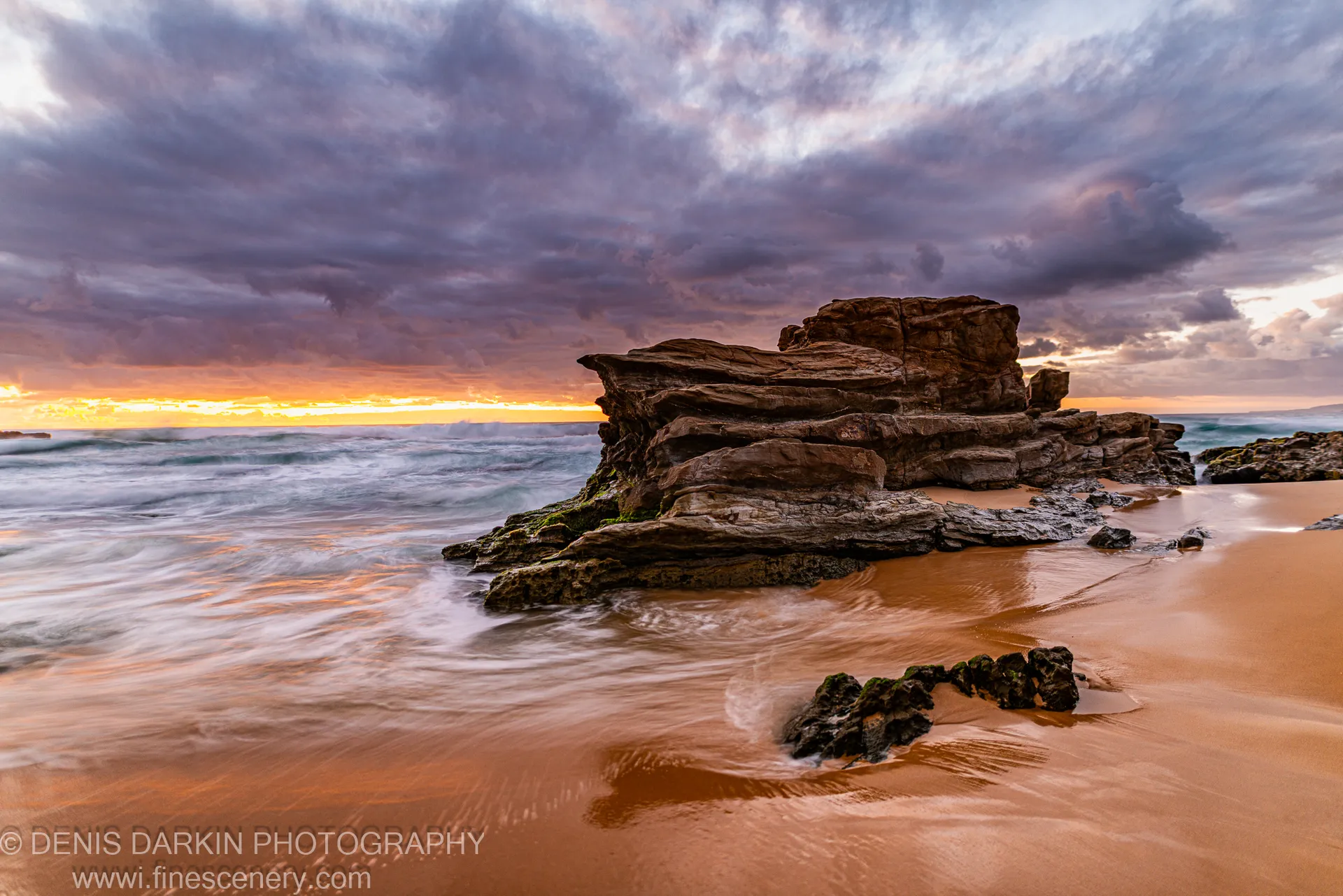 A stratified sandstone rock stack on the South Coast at dawn, with storm clouds above and long-exposure surf on wet sand.