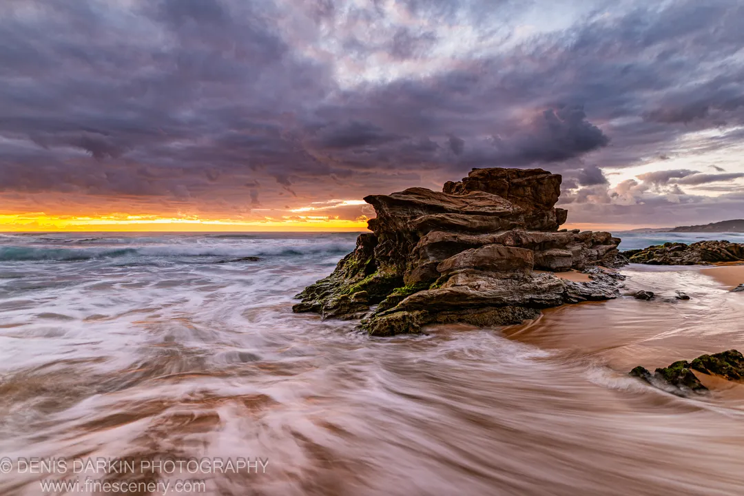 Long-exposure ocean foam washing over moss-covered sandstone rocks on the New South Wales South Coast at sunrise, with storm clouds above a bright horizon.