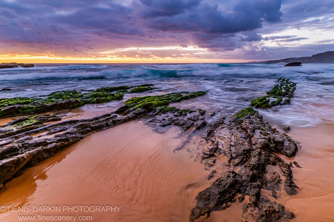Moss-covered rock formations extend across a wet sandy beach on the South Coast at dawn, with motion-blurred surf and storm clouds gathering overhead.