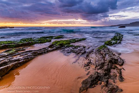 Moss-covered rock formations extend across a wet sandy beach on the South Coast at dawn, with motion-blurred surf and storm clouds gathering overhead.