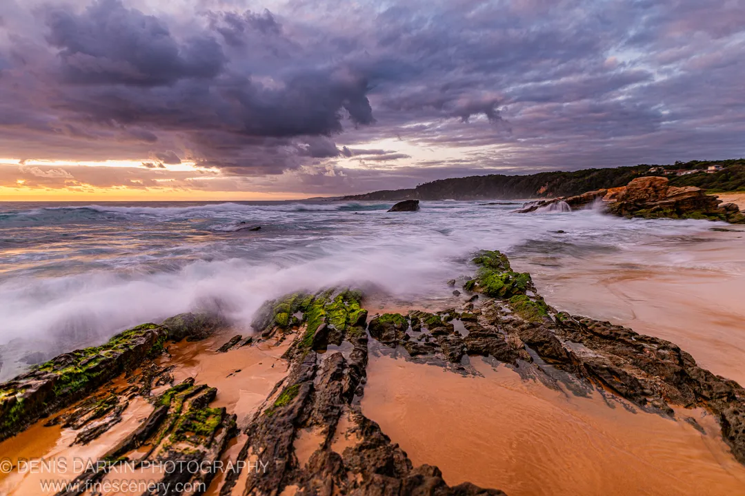 Algae-covered rock formations stretch toward the ocean on the South Coast at dawn, with storm clouds above a golden horizon.