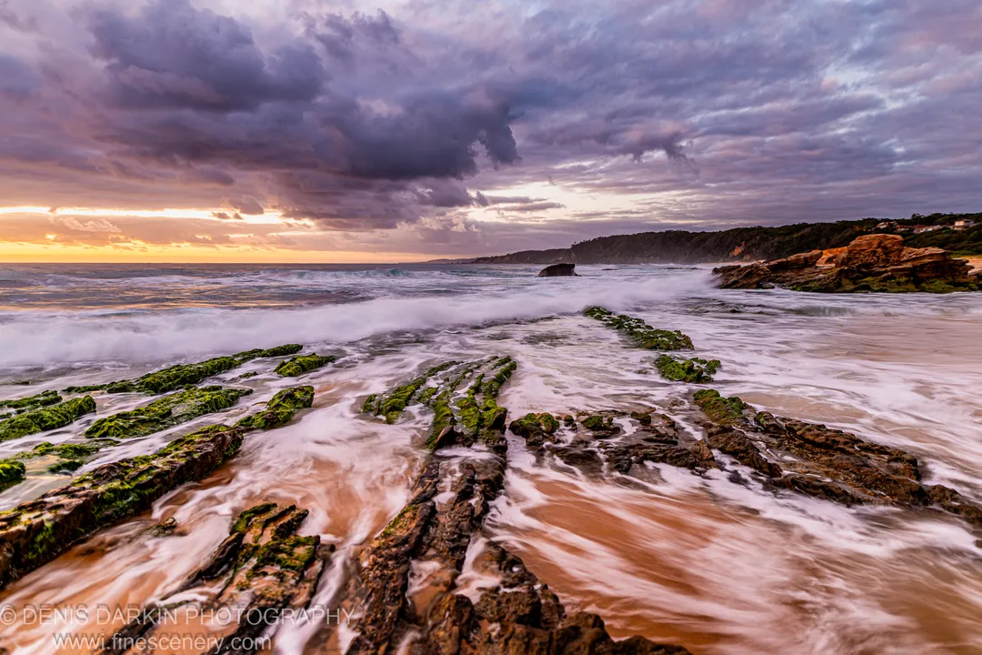 Water rushes in a long-exposure surge over an algae-covered rock platform on the South Coast at sunrise, with storm clouds and warm light overhead.