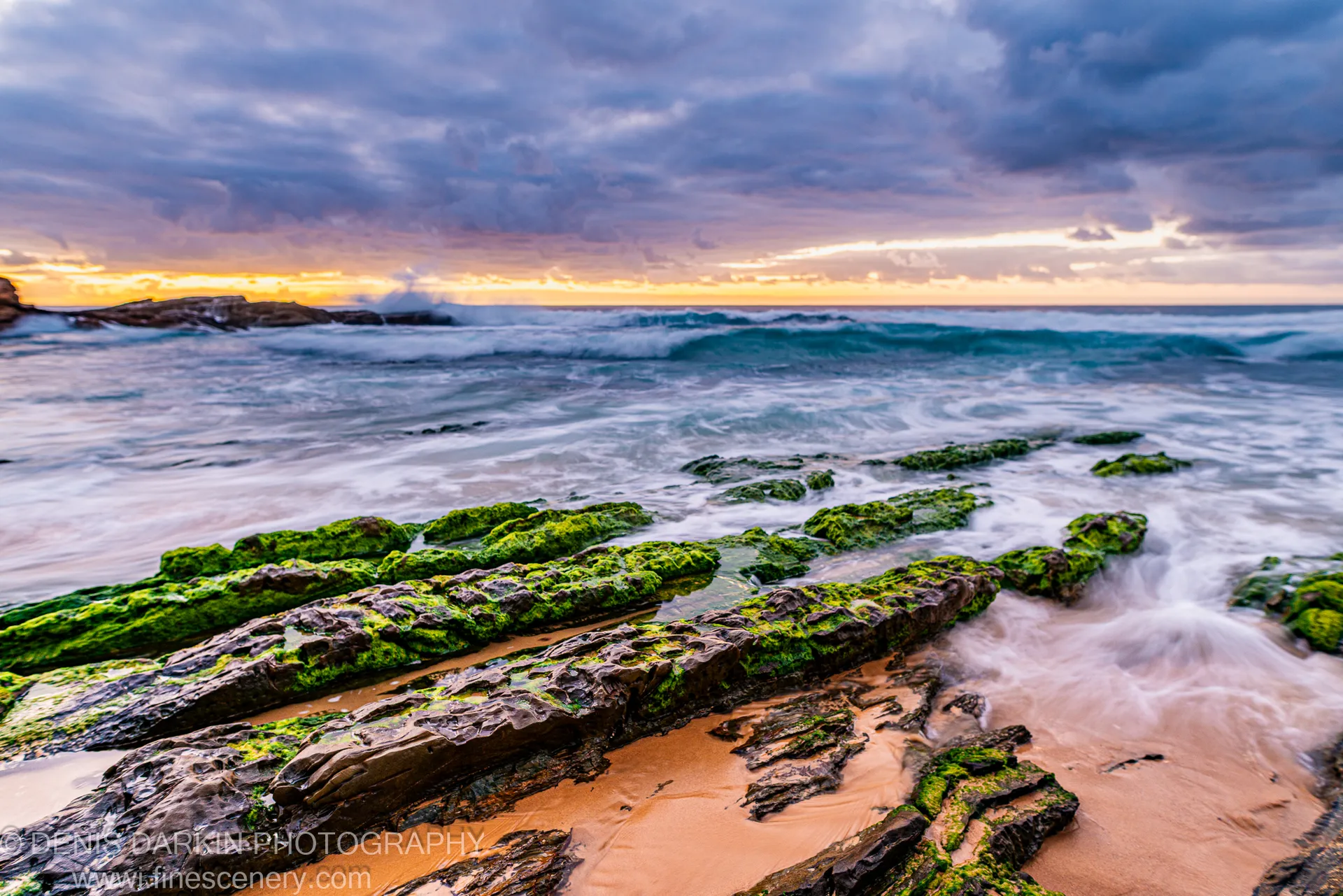 Long-exposure surf rushes over moss-covered rock shelves on the South Coast at dawn, with a breaking wave in the distance under a heavy storm sky.