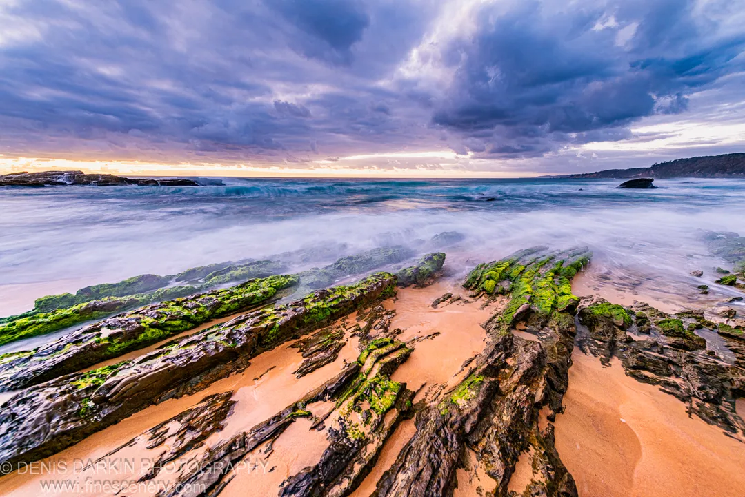 Algae-covered rocks cut through wet sand on the South Coast at dawn, with long-exposure surf and a coastal headland visible under heavy storm clouds.