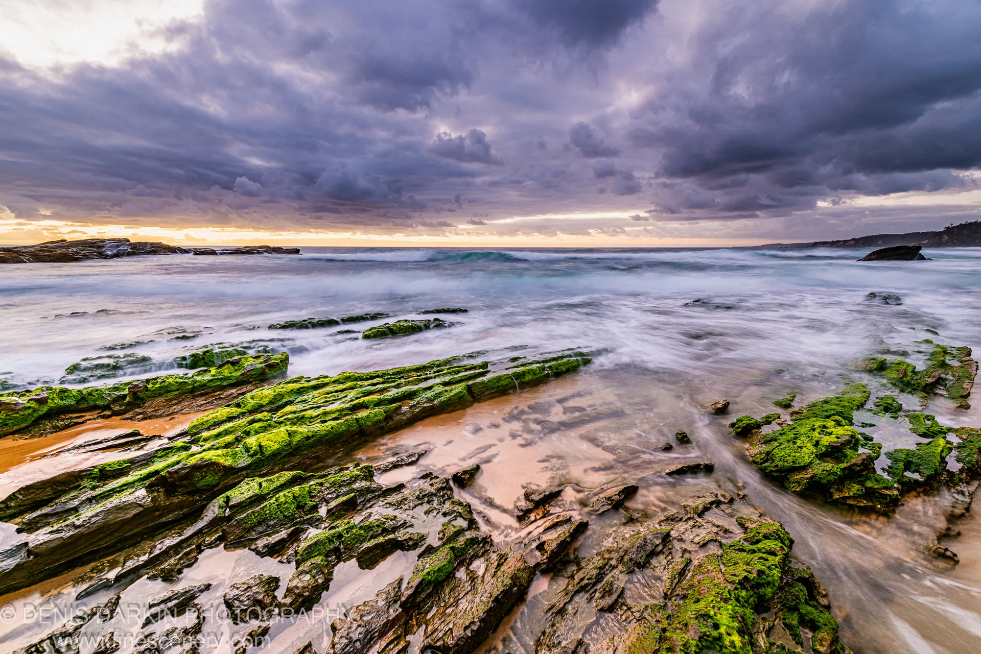 Green algae covers the stratified rock platform in the foreground on the South Coast at dawn, with long-exposure surf and heavy storm clouds overhead.