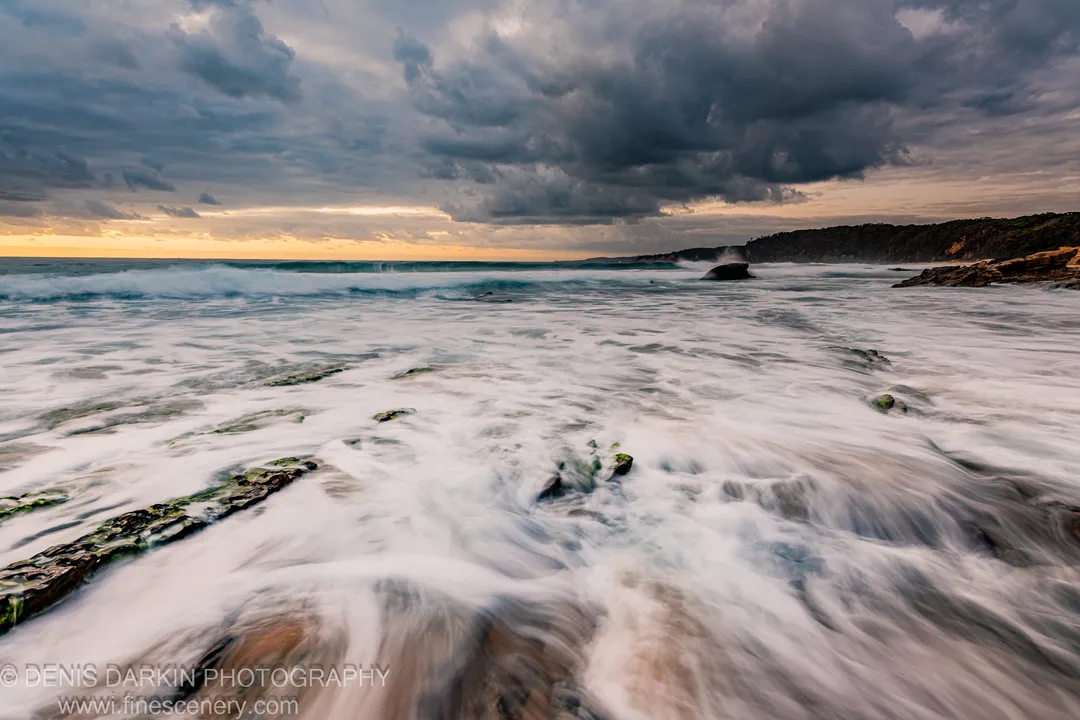 Long-exposure surf over moss-covered rocks at dawn on the New South Wales coast, heavy storm clouds above a warm golden horizon strip.